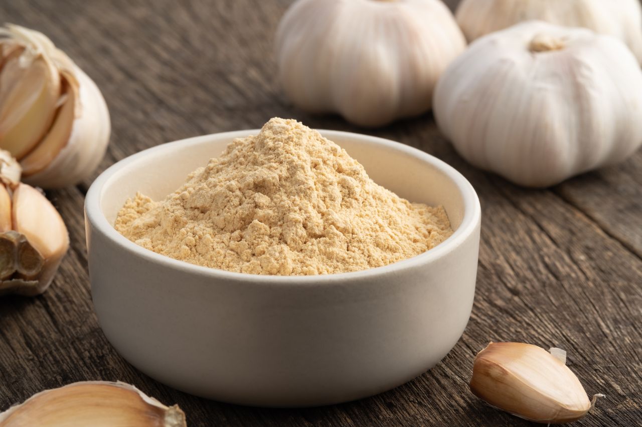 A bowl of garlic powder surrounded by garlic cloves and bulbs on a wooden surface