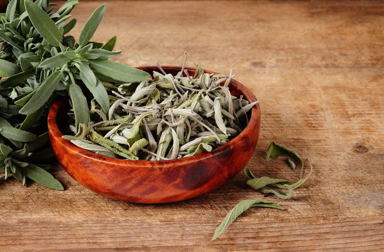 A wooden bowl containing dried sage leaves alongside a bundle of fresh sage on a wooden surface