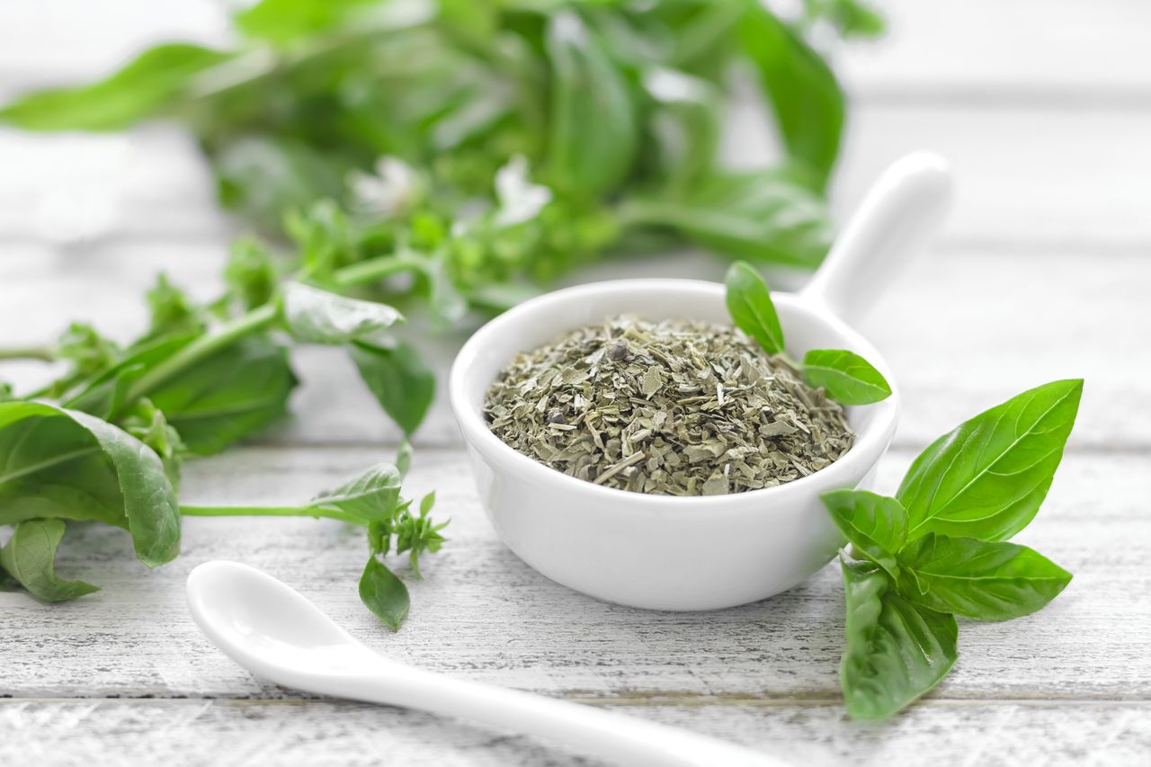 A bowl of dried herbs surrounded by fresh green herb leaves on a wooden surface
