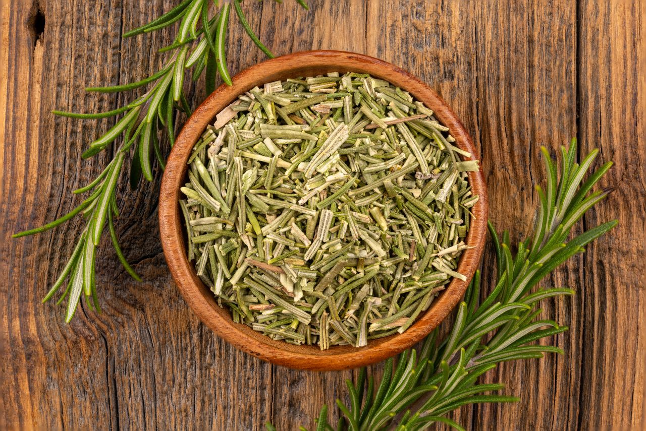 Dried rosemary in a wooden bowl on a rustic wooden surface with sprigs of fresh rosemary