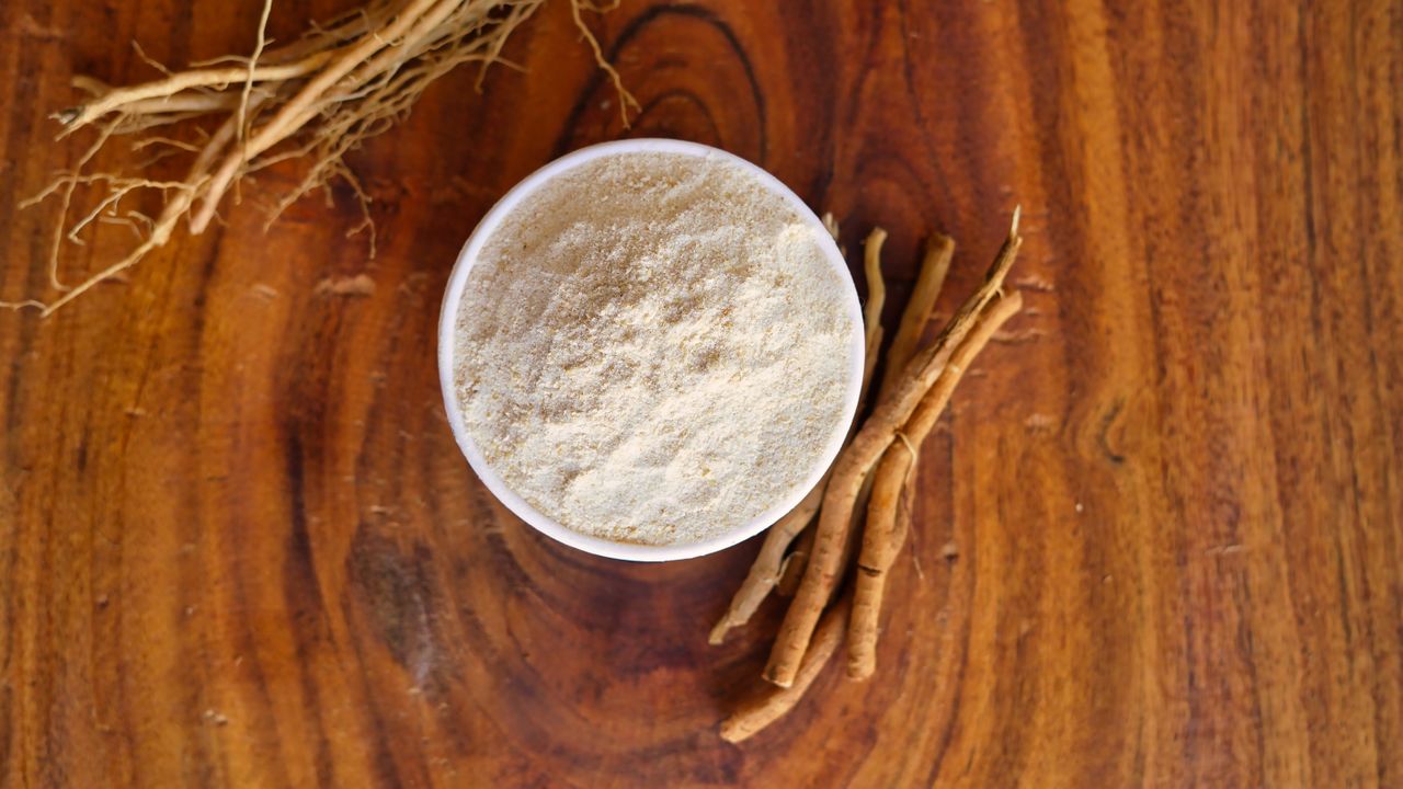 A bowl of powdered ashwagandha root with roots beside it on a wooden surface