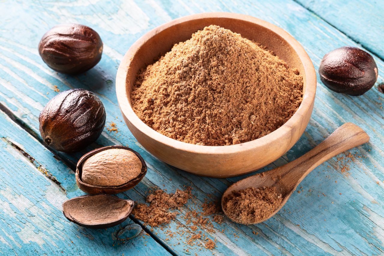 A bowl of ground nutmeg surrounded by whole nutmeg seeds and pieces on a wooden surface