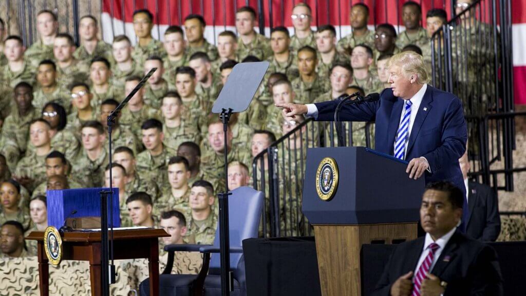 President Donald Trump speaking at a podium with military officials nearby.