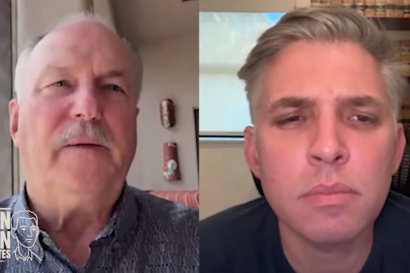 Two individuals are engaged in a discussion, likely in a professional setting. The individual on the left has a receding hairline and a mustache, wearing a light-colored shirt. The individual on the right has short, light hair, wearing a dark-colored shirt, with a serious expression. The background shows elements of an indoor setting, possibly an office or a home office.