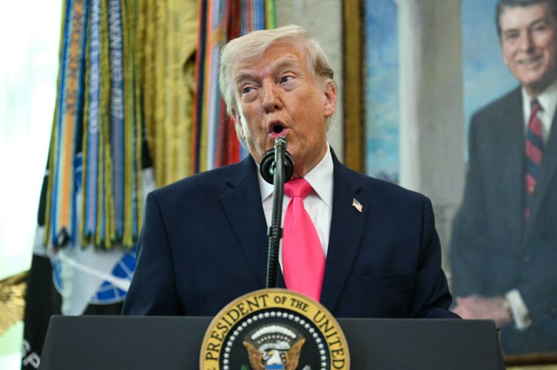A person in formal attire, speaking into a microphone at a podium adorned with the Seal of the President of the United States, with a framed portrait of another individual in a suit and tie in the background.
