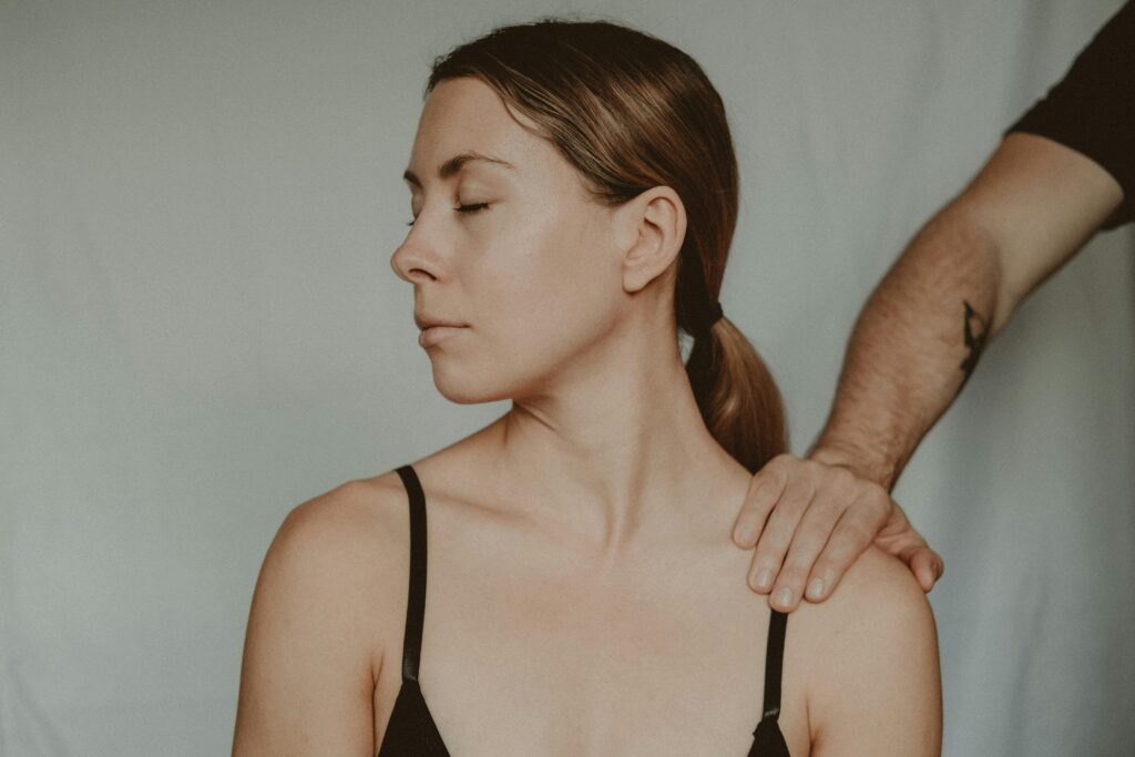 mage 12: Woman with reddish-brown hair in a low ponytail, eyes closed, wearing a black tank top. A hand rests on her shoulder.