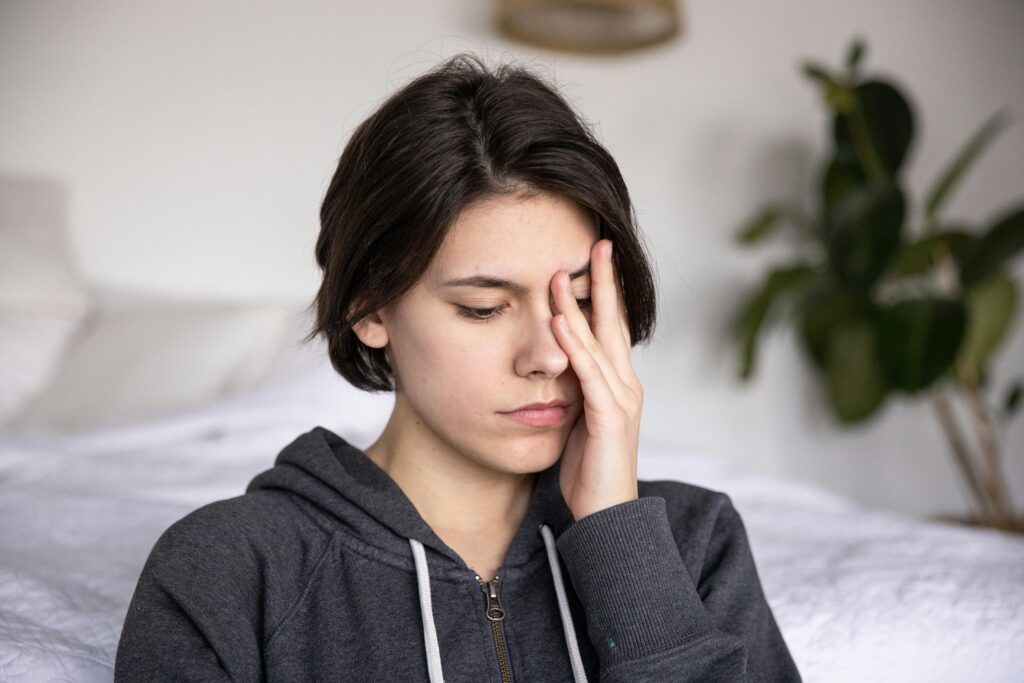 Young person with short dark hair sits looking tired or stressed, rubbing their eye with one hand. They wear a grey zip-up hoodie. A houseplant is visible in the background.
