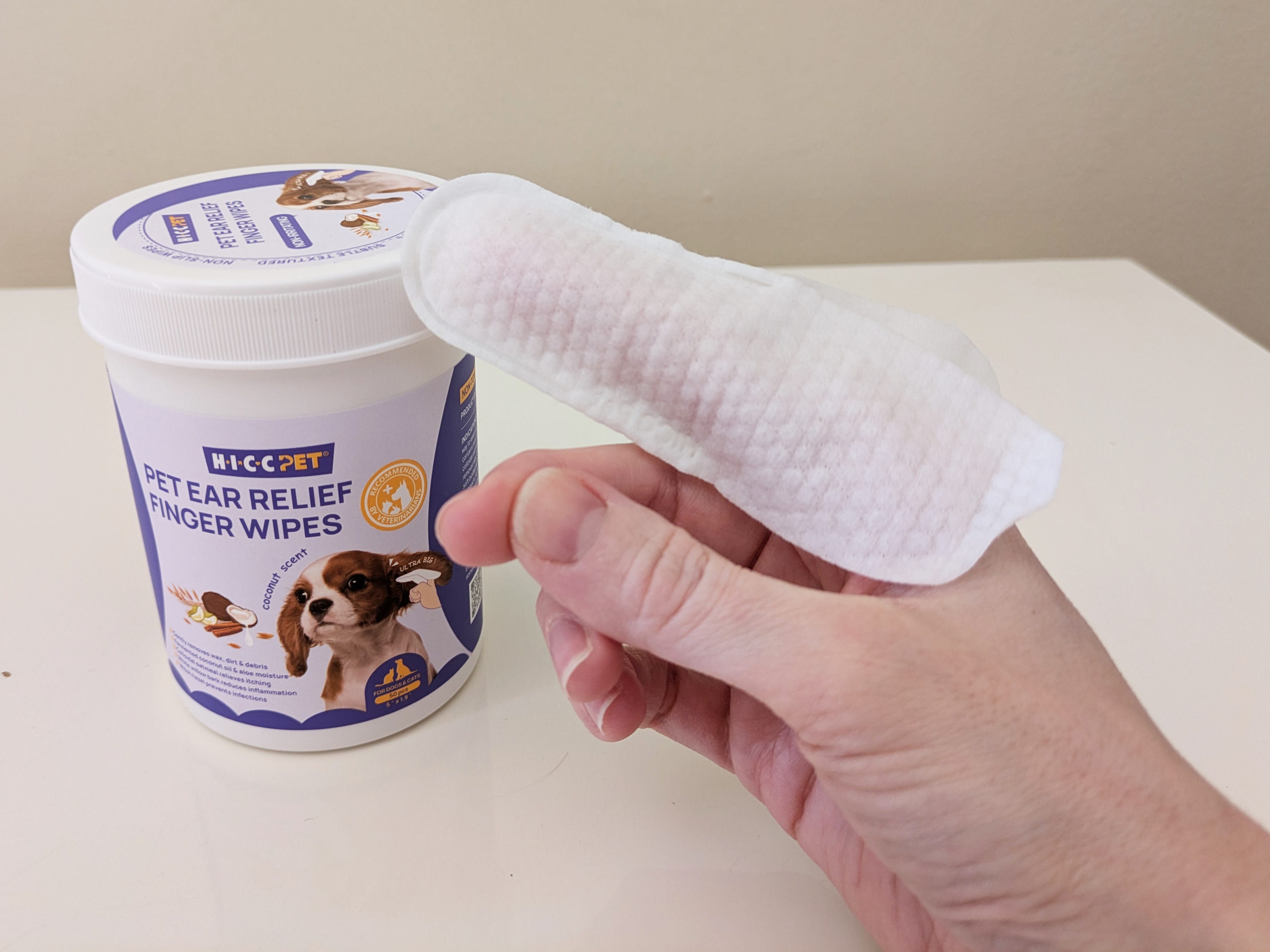 A hand holds a textured pet ear-cleaning wipe in front of a container of HICC Pet Ear Relief Finger Wipes on a white table.