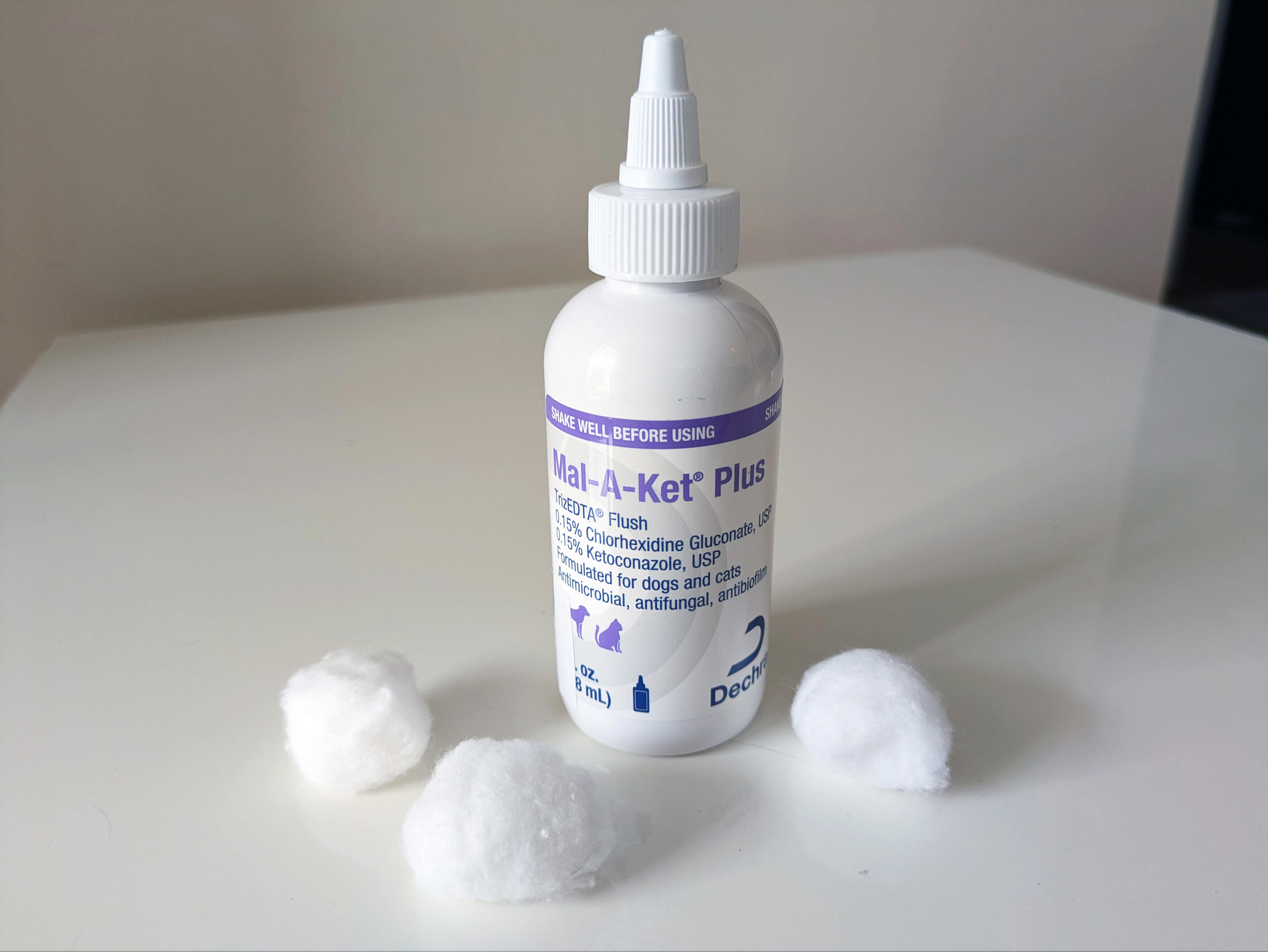 A bottle of medicated Mal-A-Ket pet ear-cleaning solution stands on a white surface with three cotton balls placed around it.