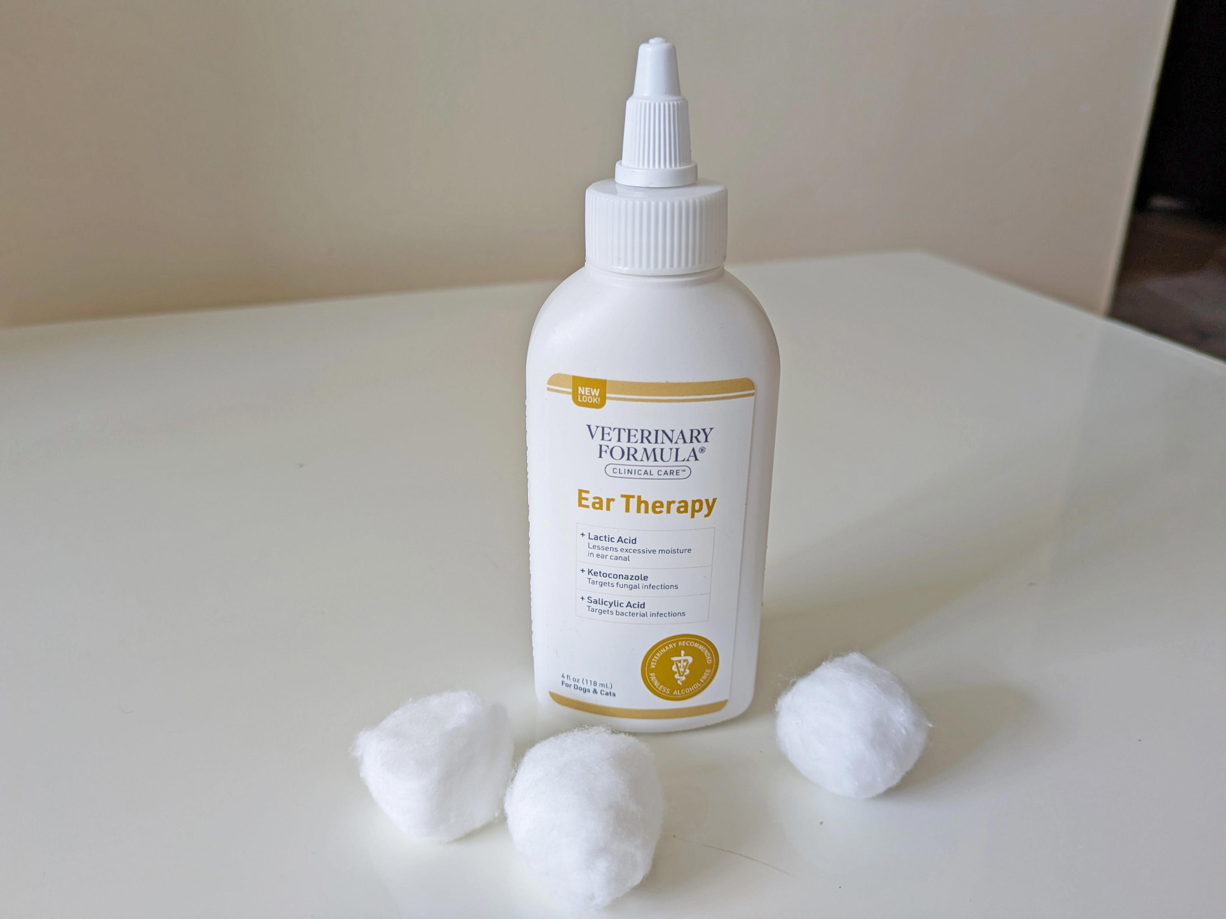 A bottle of Veterinary Formula ear-cleaning solution stands on a white table with three cotton balls placed in front of it.