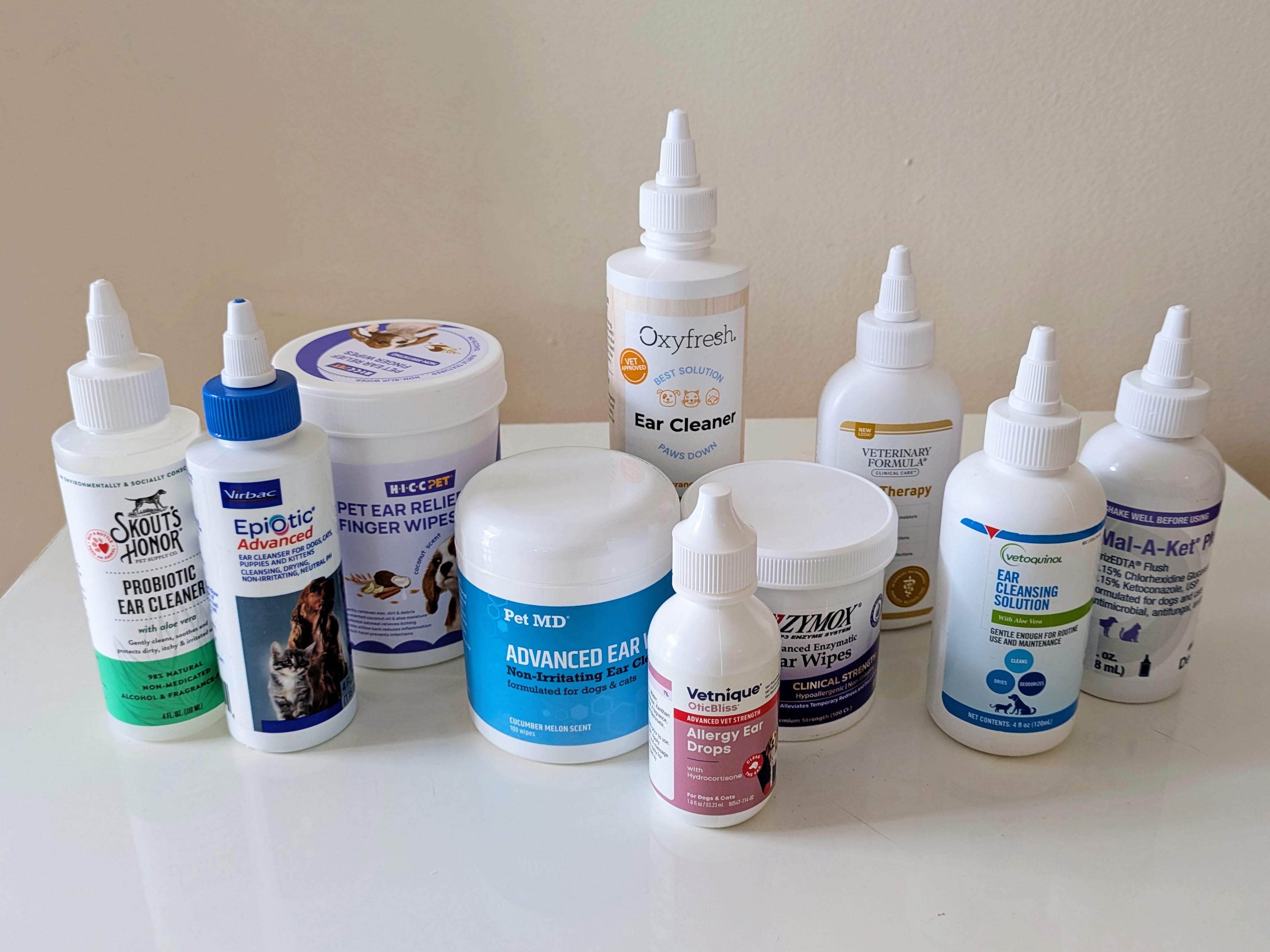 A variety of dog ear-cleaning products, including solutions, wipes, and drops, are arranged on a white table against a plain background.