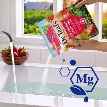 Bag of Dr. Teal's Epsom salt with strawberry scent is poured into a bathtub. A bowl of strawberries is on the edge of the tub