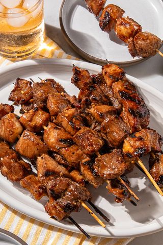 Grilled pork skewers on a white platter with a drink in the background