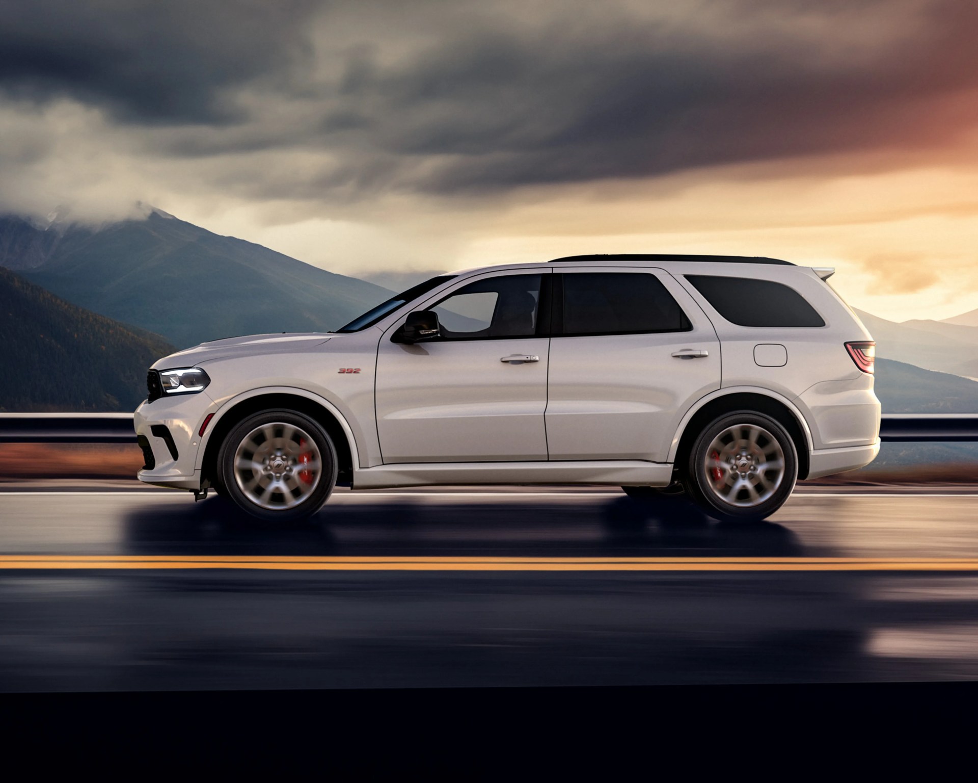White Dodge Durango SUV with red brake calipers driving on a road with mountains and cloudy sky in the background.