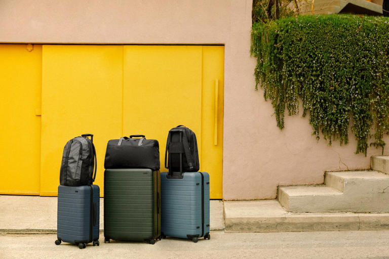 Away Luggage Set with Away bags on top, against a tan wall with tan stairs on the right and yellow panels on the left