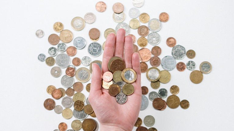 Eine Person hält Münzen verschiedener Währungen in einer Hand, im Hintergrund noch mehr Geld