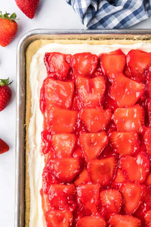 A close up of the corner of a sheet pan full of strawberry pizza dessert with glazed strawberries on top of a frosting and a sugar cookie base on a counter.