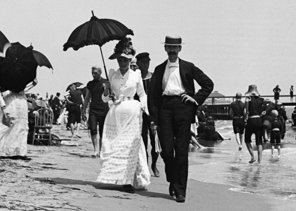 <p>Seaside socializing rose to popularity in the 19th century, beginning in Britain where doctors were <a href="https://www.denverpost.com/2016/07/07/origins-of-beach-vacations/">encouraging beach visits as a way to combat "melancholy,"</a> and later in America as the trend hit the East Coast. The <a href="https://web.archive.org/web/20220202074532/https://archive.curbed.com/2018/7/19/17590246/boardwalk-history-coney-island-atlantic-city">first boardwalk stateside was built in Atlantic City</a> in 1870 by two men—a hotelier and a railroad conductor—who had grown aggravated with beachgoers consistently dragging sand into their resorts and train cars. It was very basic, consisting only of an arrangement of boards laid out on the sand, and was later replaced by a larger railed boardwalk in 1890. In this photo, people stroll along the sand at the New Jersey beach, with the boardwalk in the background.</p>