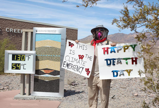 A climate change protestor stands next to a digital display of an unofficial heat reading at Furnace Creek Visitor Center during a heat wave in Death Valley National Park in Death Valley, California, on July 16, 2023.