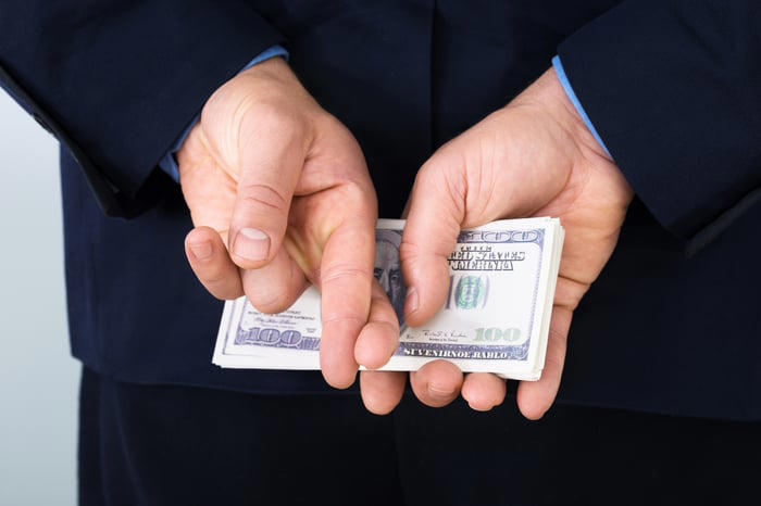 A businessperson crossing their fingers behind their back while holding a stack of one hundred dollar bills. 