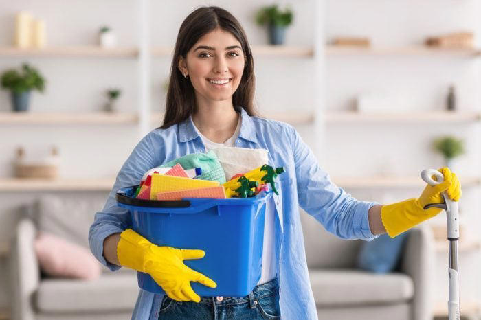 Portrait of smiling millennial cleaning lady holding bucket with cleaning supplies and mop, posing and looking at camera standing in living room