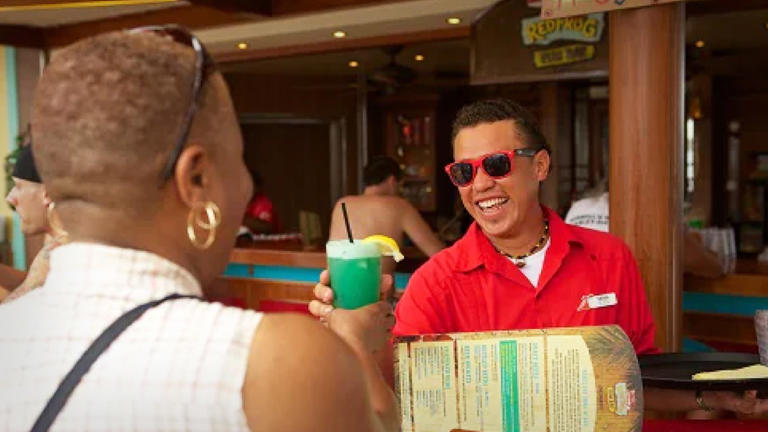 A Carnival Cruise Line bartender hands a passenger a drink. Bar Lead JS