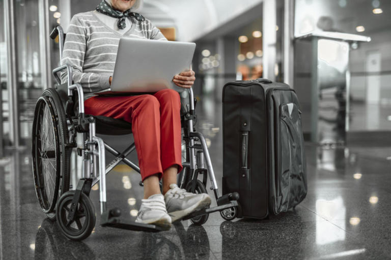 Woman sitting in a wheelchair at the airport, with a laptop in her lap and a suitcase beside her.