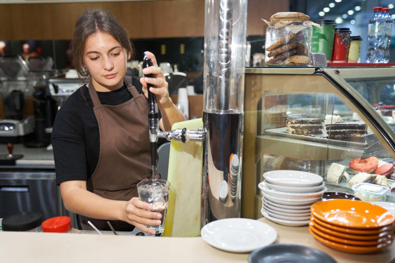 A young beautiful woman in a brown apron is standing and pouring water into a glass. The concept of a small business. Work for yourself.