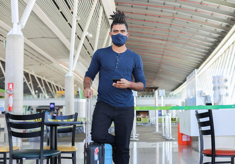 12 travel essentials for men that are actually useful and will make traveling easier. Pictured: a man wearing a mask standing in front of the camera with a suitcase.
