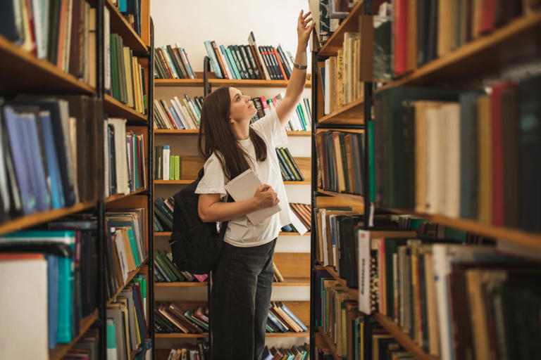 Beautiful female student standing in university library, looking for a book and searching information for a project, learning for passing exams, education concept.