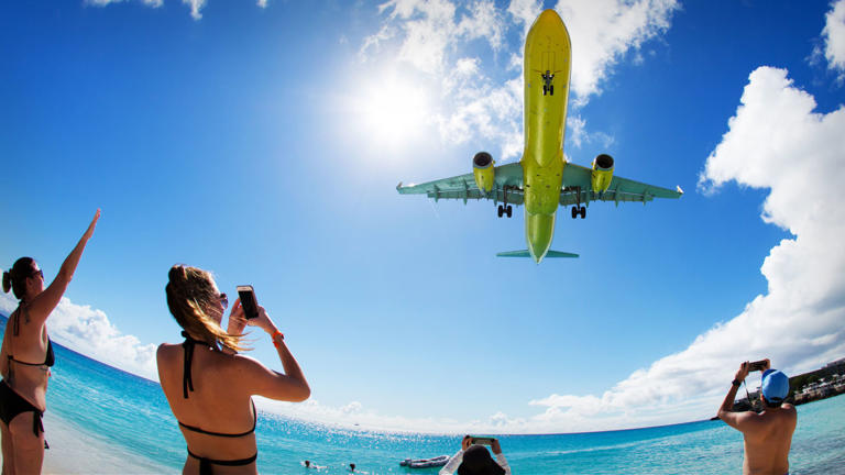 A Spirit Airlines plane is seen flying over onlookers taking photos on a beach. -lead