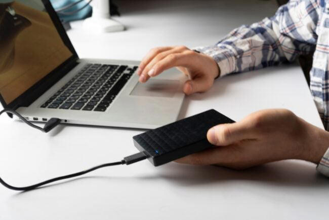 A young holding an external hard drive that is plugged into a laptop.