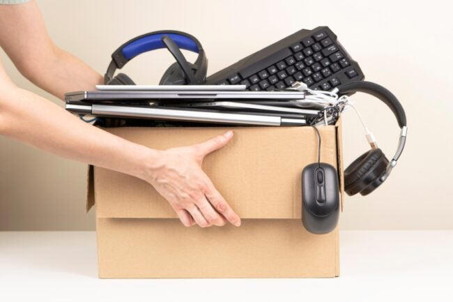 A person lifting a box full of laptop, computer peripherals, and pairs of headphones.