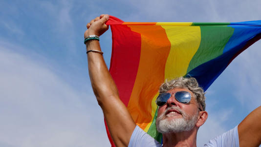 An older man with sunglasses waving a rainbow flag.