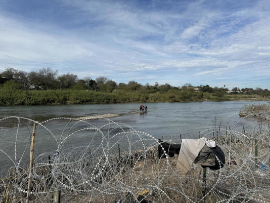 A group of migrants wait in the middle of the Rio Grande near Eagle Pass, looking at a U.S. border fortified by coils of concertina wire and Texas National Guard troops. / Credit: Camilo Montoya-Galvez
