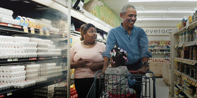 Barack Obama is pushing a child in a grocery cart while her mom watches. 