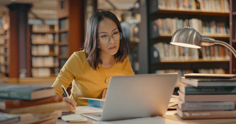 Lawyer doing research on the internet using her laptop. Young woman working on a case. Female alone inside a library working on a project