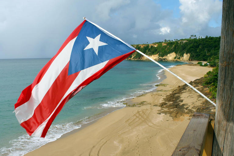 Puerto Rico is a safe island to visit in the Caribbean. Learn more about any possible threats to tourists and what they can do to ensure their safety. pictured: a Puerto Rican flag posted off of a building on the sandy beach during a sunny day