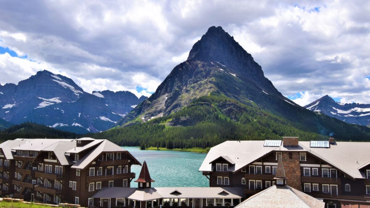 The historic Many Glacier Hotel with its distinctive alpine architecture, overlooking the turquoise waters of Swiftcurrent Lake and flanked by towering, rugged mountain peaks under a cloudy sky.