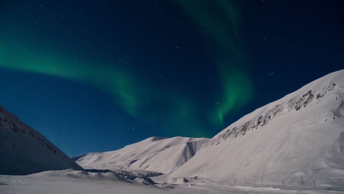 he skies over Svalbard are painted with a brilliant display of green and purple auroras, shining over a vast snowfield with dark, rugged mountains providing a dramatic contrast.