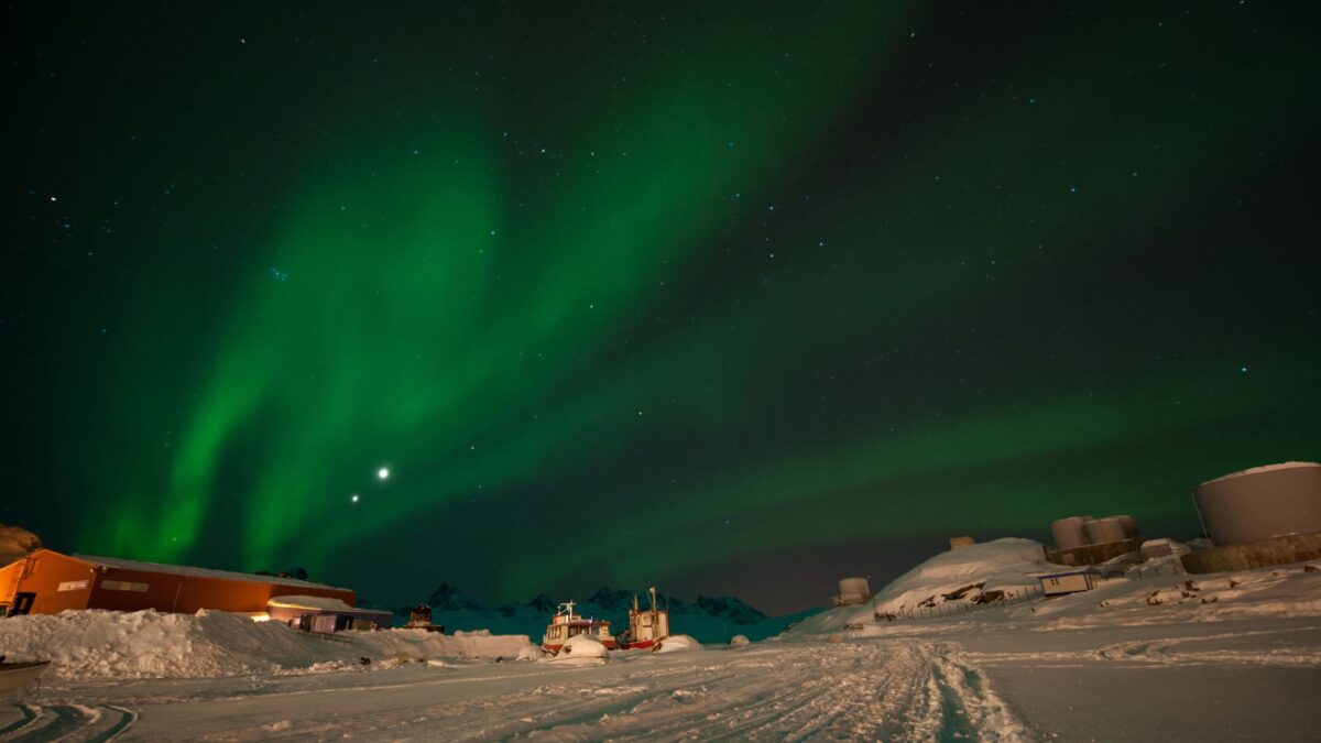 The aurora borealis dances above a small Greenlandic settlement, casting a green light over the snow-covered houses and the icy terrain, with a backdrop of dark, starry skies.