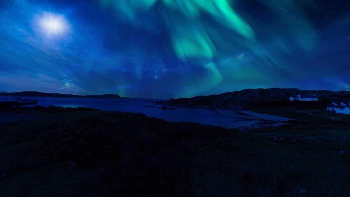 he Northern Lights appear as a celestial dance of vibrant greens and blues above a rugged Scottish coastline. The moon adds a bright focal point amidst the aurora's glow, while the silhouetted terrain and houses add a sense of solitude and wonder