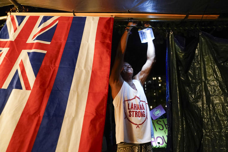 FILE - Naldo Valentine, who lost his home to the Lahaina wildfire, puts up a light as darkness falls at a housing protest on Kaanapali Beach Wednesday, Dec. 6, 2023, in Lahaina, Hawaii. A group of survivors were camping on the resort beach to protest and raise awareness for better long-term housing options for those displaced. (AP Photo/Lindsey Wasson, File)