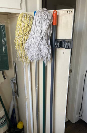 Three mops, one yellow and two white, hang on a utility rack next to a broom with a red handle in a storage area. Cleaning supplies are visible on the left