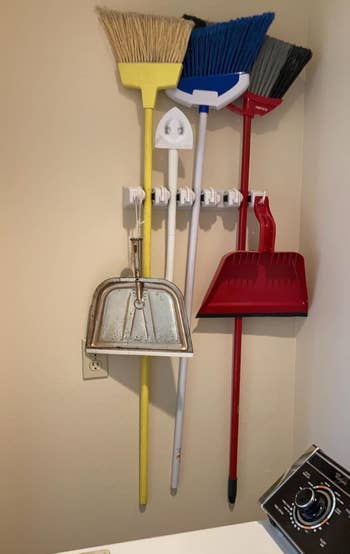 Various cleaning tools including brooms, dustpans, and a mop are hung on wall hooks in a laundry room. A washing machine corner is visible