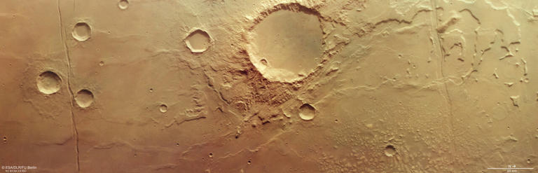 This tan-colored slice of ground is smoother to the left and becomes covered in small hills and mounds to the right. Wriggly, uneven ridges cut horizontally across the frame, while two prominent fault lines cut down vertically on the left and right. Large and small craters are peppered across the terrain. The worn-away boundaries of a once-colossal lakebed can be seen curving up and away from the bottom-center to the top right; this skirts around the largest crater seen here, which sits in the middle of the picture encircled by rough, irregular valleys and channels. Credit: ESA/DLR/FU Berlin