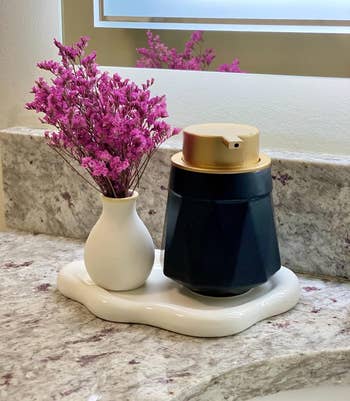 Purple flowers in a white vase next to a black and gold soap dispenser on a marbled countertop