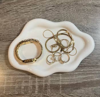 A white dish holds a gold watch, three gold rings, and a tangled gold necklace on a wood-textured surface
