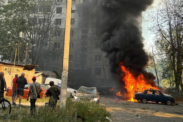 Local residents stand next to a burning car in the courtyard of a damaged residential building following a missile attack in Kharkiv