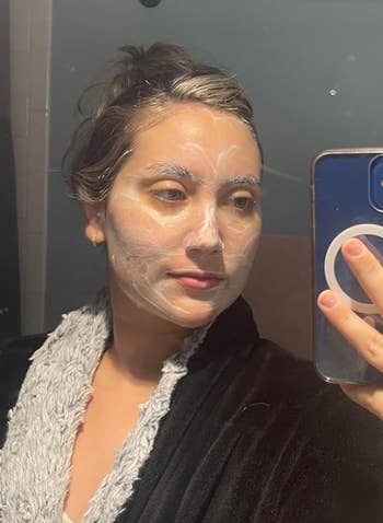 Person taking a mirror selfie while wearing a facial mask, dressed in a cozy robe with a fluffy collar